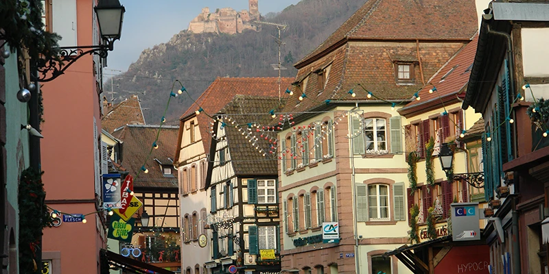 Colorful houses along a Ribeauvillé street with a castle ruin on the hillside above