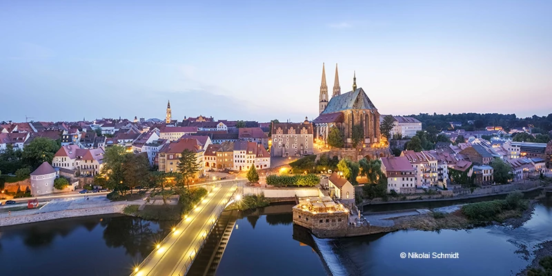 Görlitz old town skyline at dusk seen across the Neisse River, with the twin spires of the Church of St. Peter and Paul lit against the sky