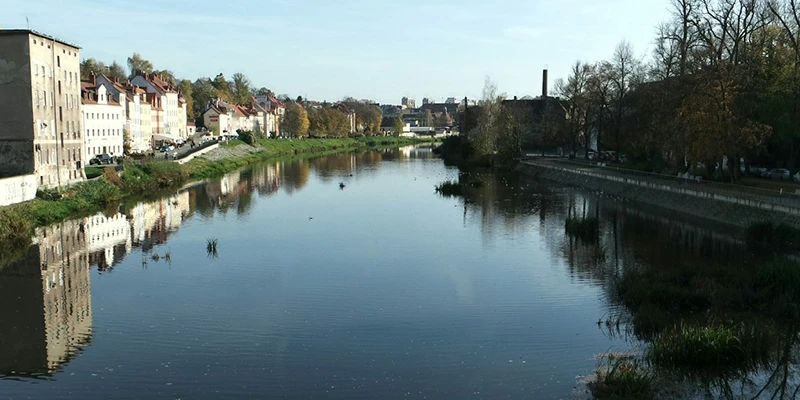 The calm Neisse River reflecting riverside houses and trees in Görlitz, with the town rising on the left bank under a clear sky