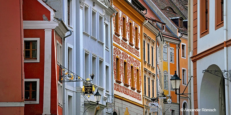 A narrow cobbled lane in Görlitz curving between pastel-painted houses, with an arch spanning the street and a hanging lantern