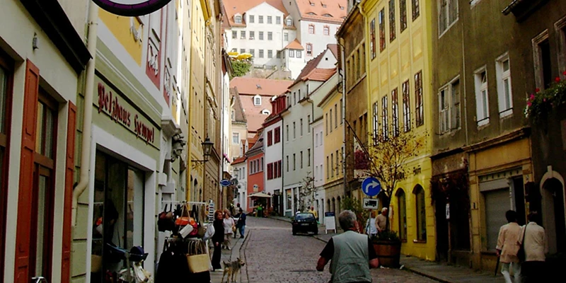 A cobbled shopping lane in Meissen climbing toward the castle, with people and a dog
