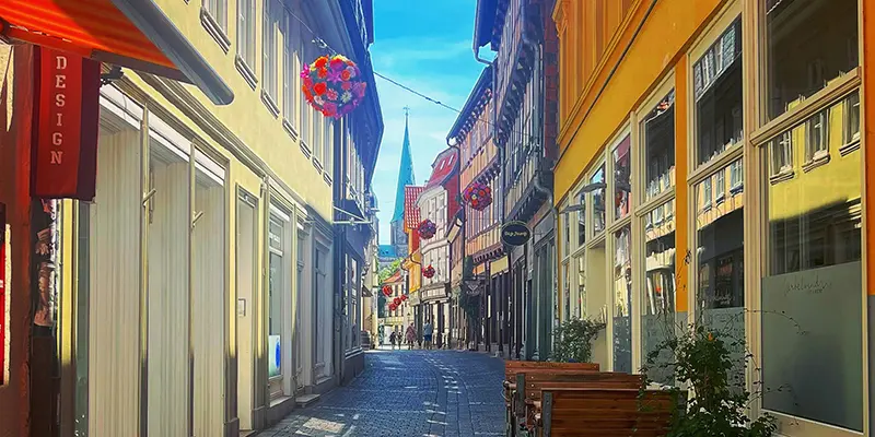 A sunlit pedestrian shopping lane with flower baskets and a church spire beyond