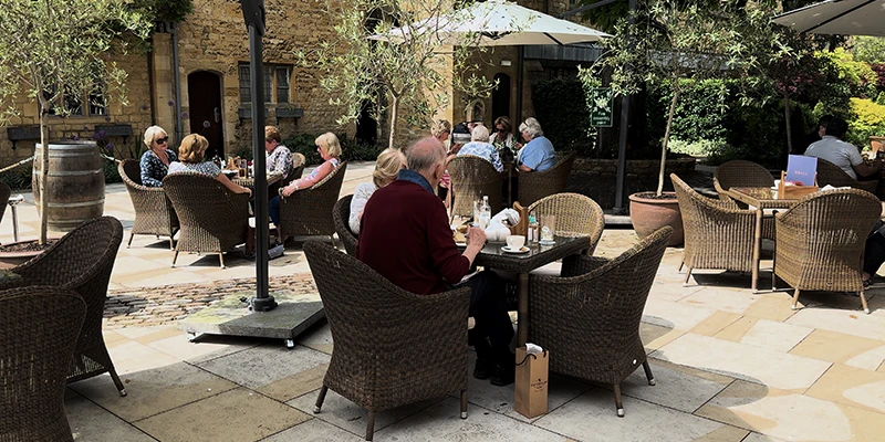 Visitors at outdoor tables in the Lygon Arms rear courtyard under cream umbrellas and a chestnut tree