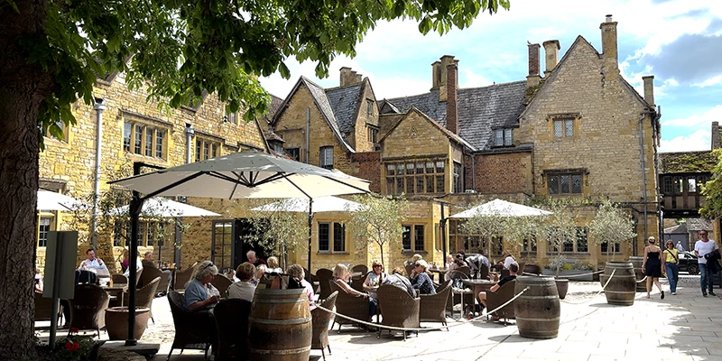 Diners enjoying lunch at outdoor tables in the Lygon Arms courtyard, with the historic honey-stone facade behind