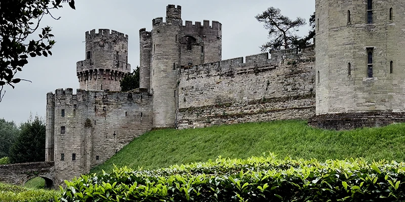 Warwick Castle seen from across the River Avon, its curtain wall and two medieval towers rising above the trees